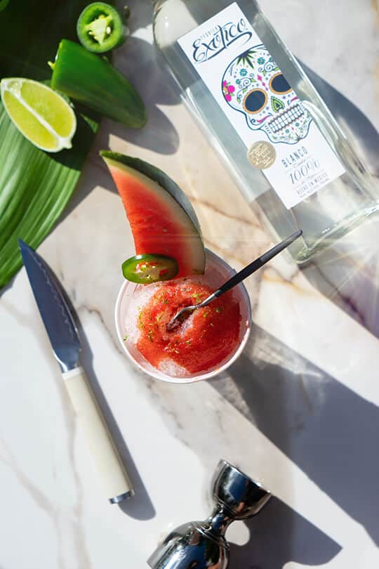 Watermelon Tequila snowcone surrounded by limes, jalapeños, a knife, shaker cup and a bottle of Exotico® Blanco.